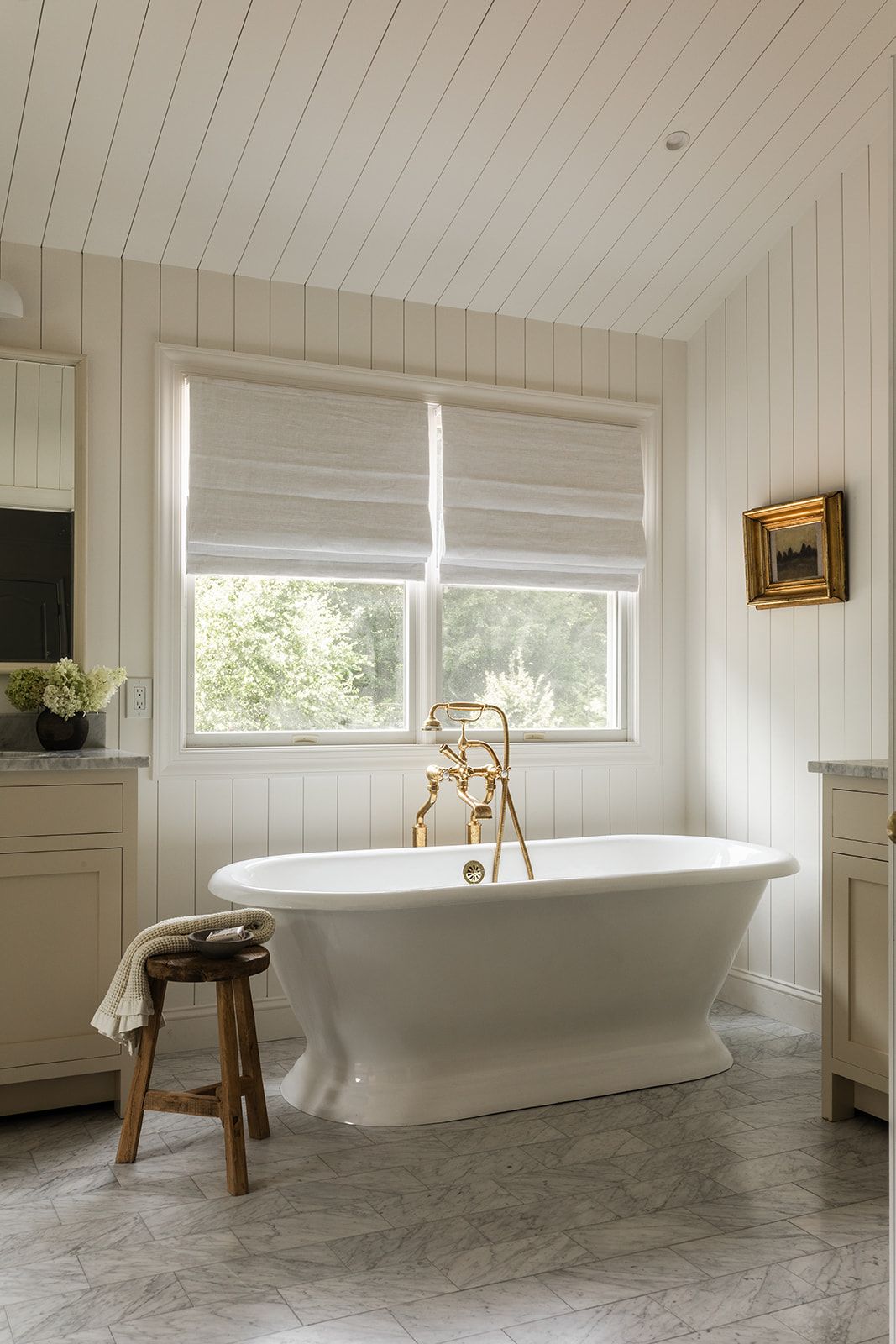 Elegant white freestanding bathtub with gold fixtures, shiplap walls, marble floors, and Roman shades in a bright bathroom.