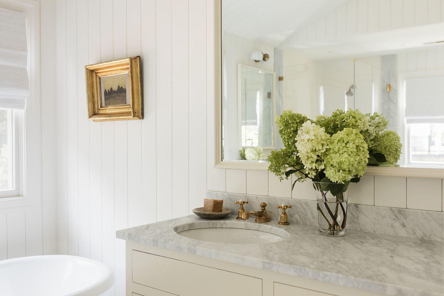 Elegant bathroom with marble countertop, white cabinetry, gold-framed mirror, and fresh green hydrangeas in a glass vase.
