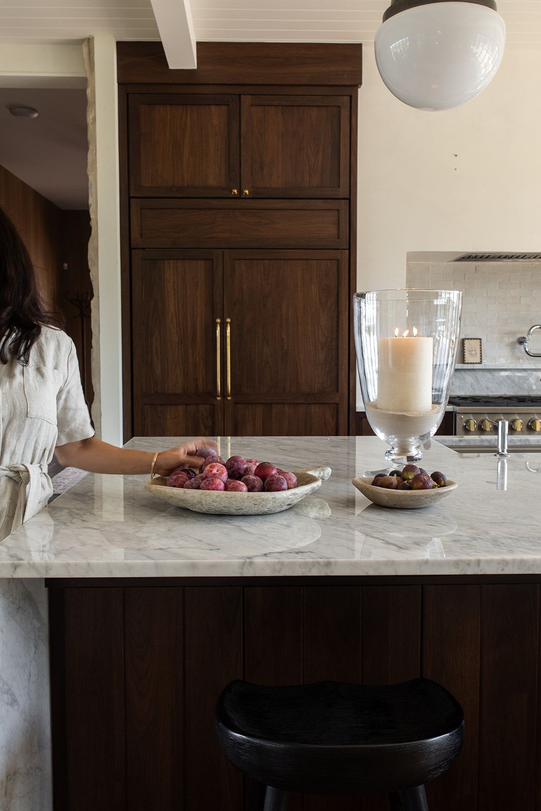 Woman in white dress at marble kitchen island with fruit bowl and candle, dark wood cabinetry with brass hardware.