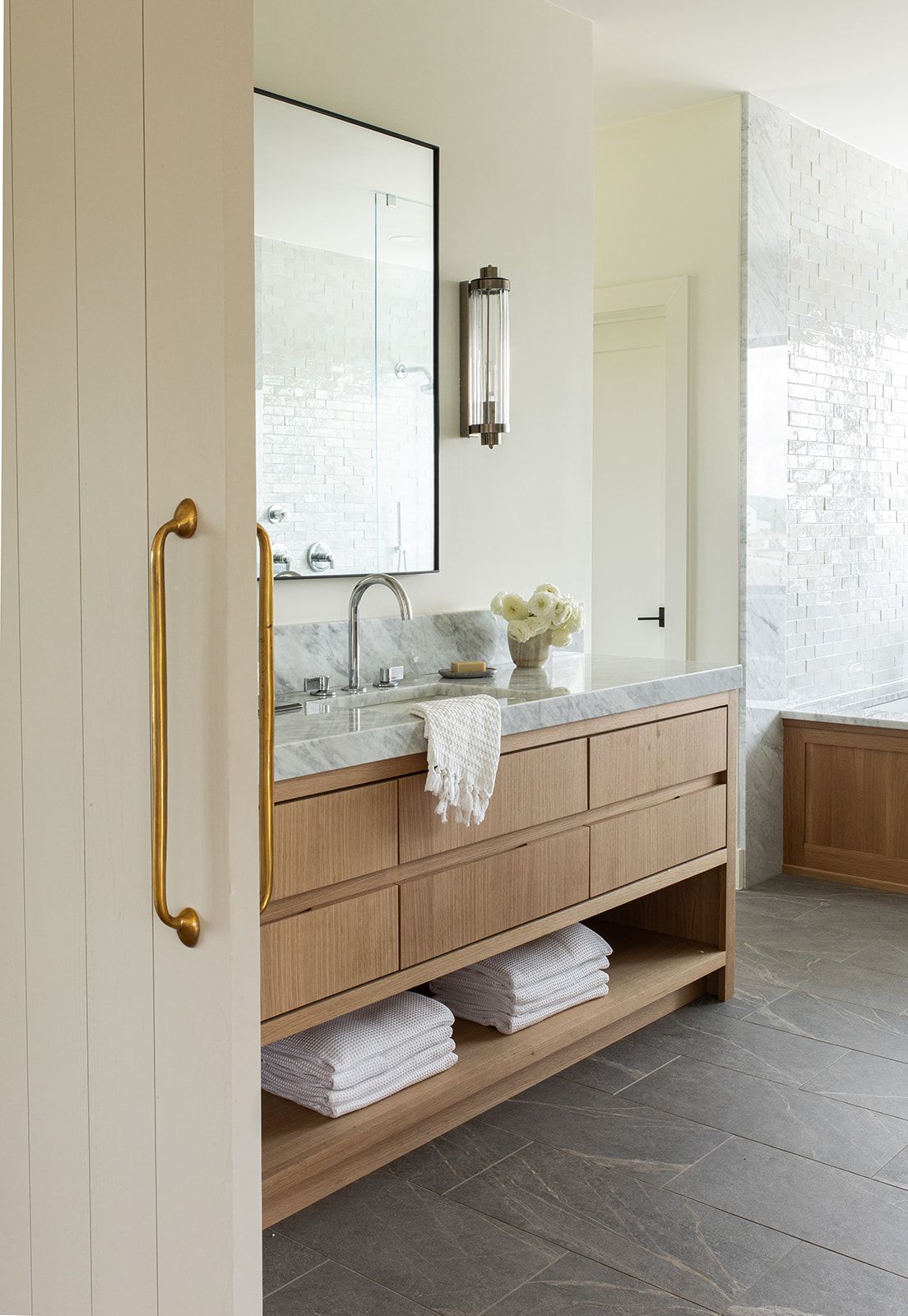 Modern bathroom vanity with light oak cabinets, marble countertop, brass pull handle, and wall sconce mirror.
