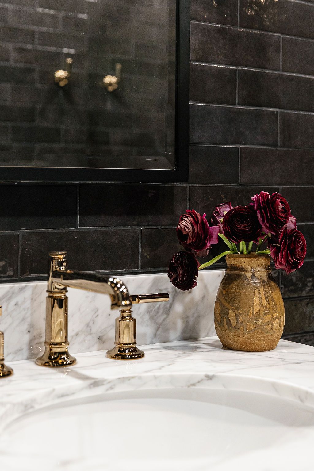 Luxury bathroom with brass faucets, dark stone wall tiles, white marble countertop, and burgundy roses in gold vases.