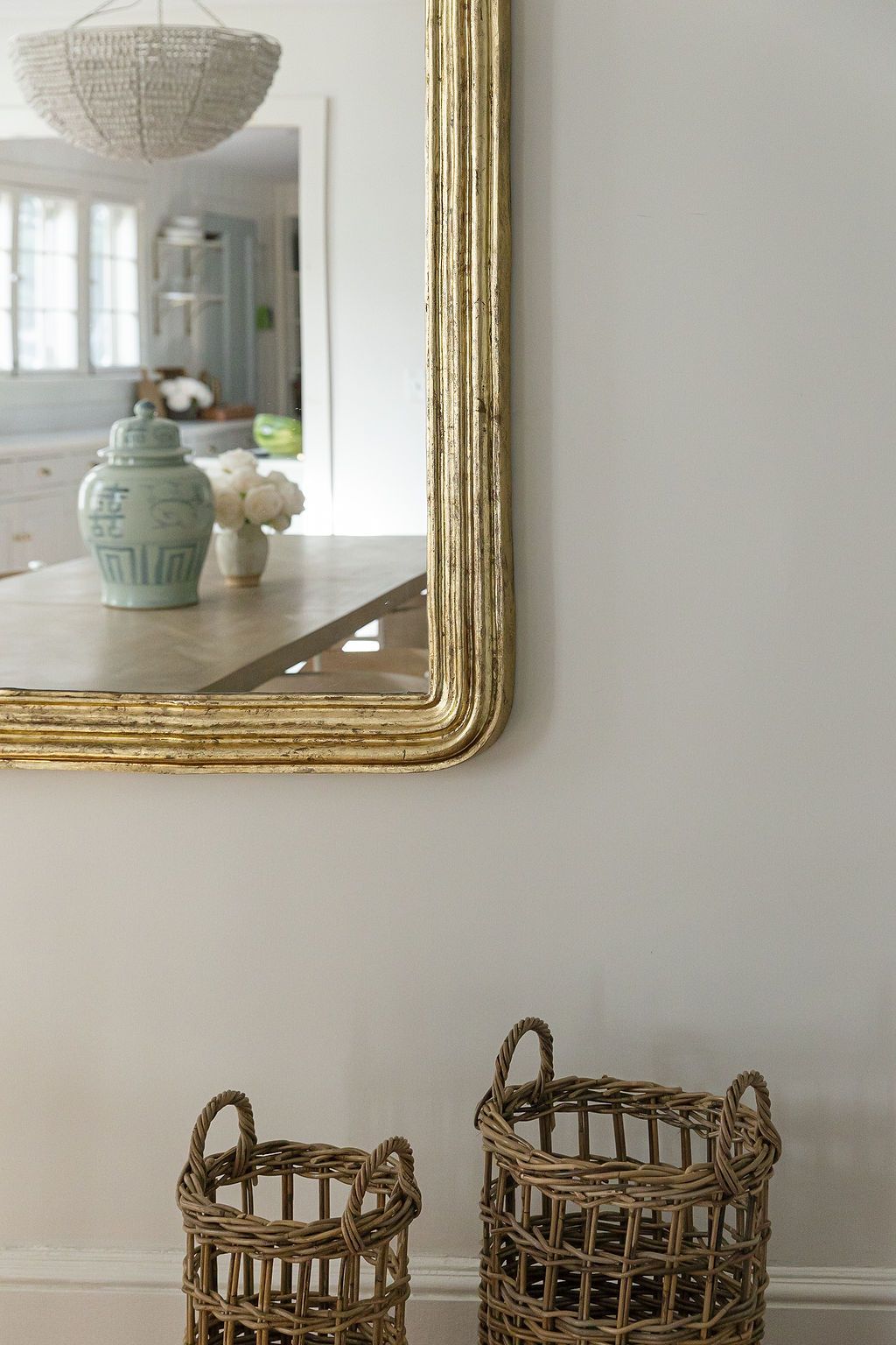 An elegant gold-framed mirror on a white wall reflects decorative objects and woven baskets in a bright interior space.