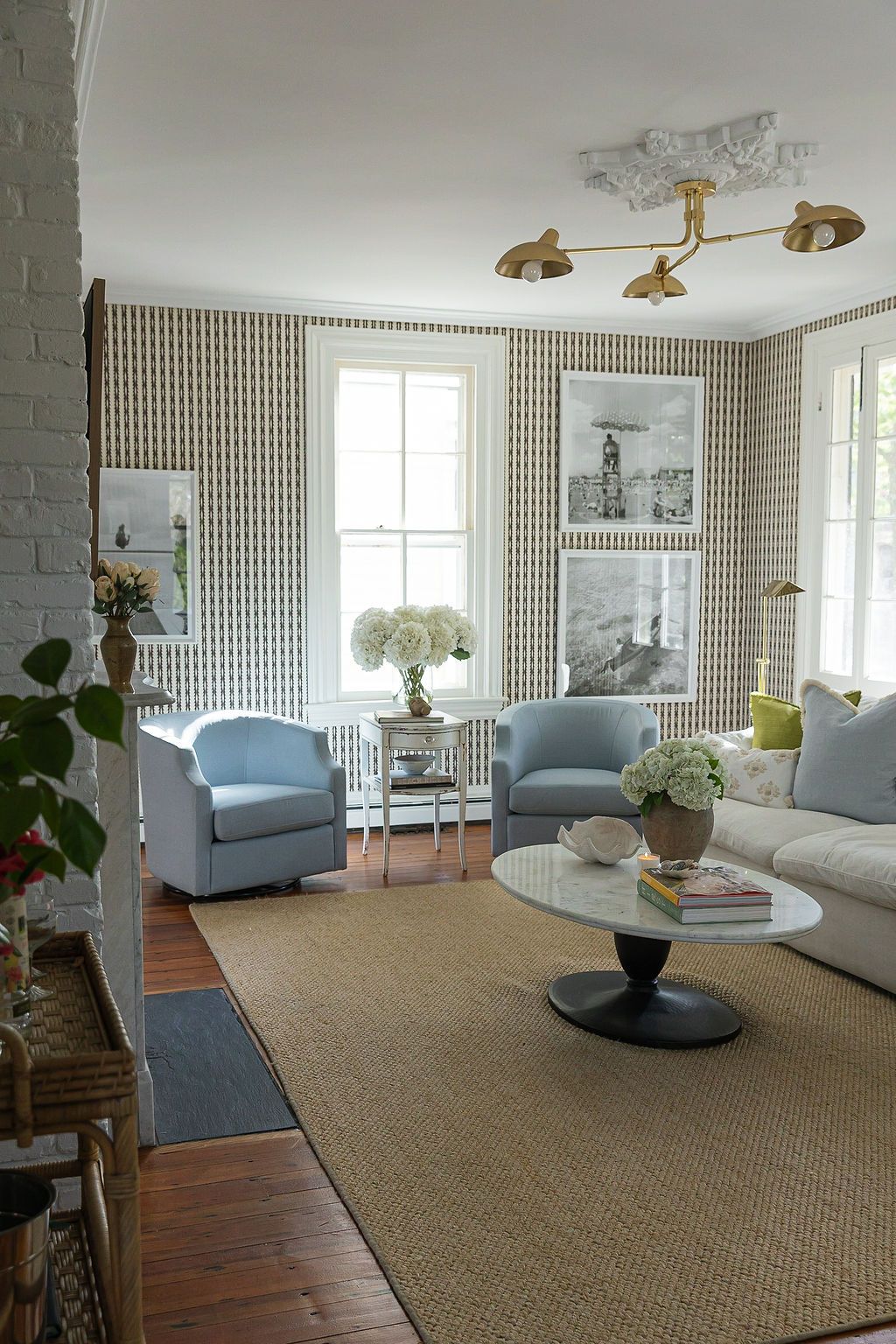 Cozy living room with blue armchairs, striped wallpaper, hardwood floors, and brass ceiling lights against natural light.