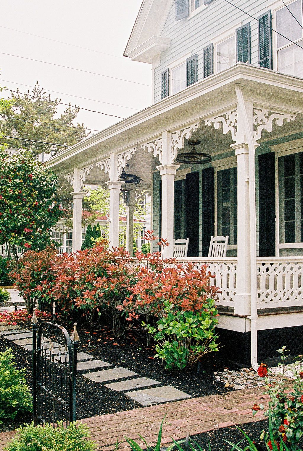 Historic white house with ornate Victorian porch features green shutters, blooming azaleas and a stepping stone path.
