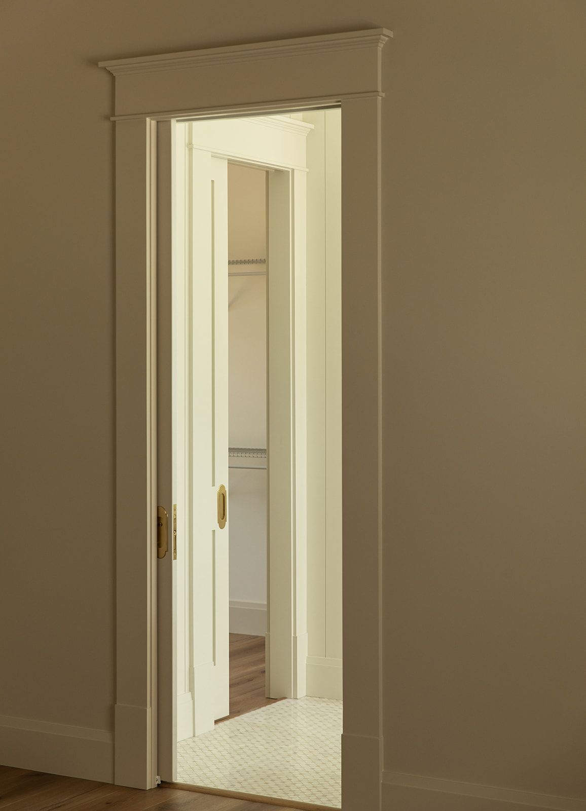 White pocket door with brass hardware opens to a bright closet, framed by elegant crown molding in a neutral room.