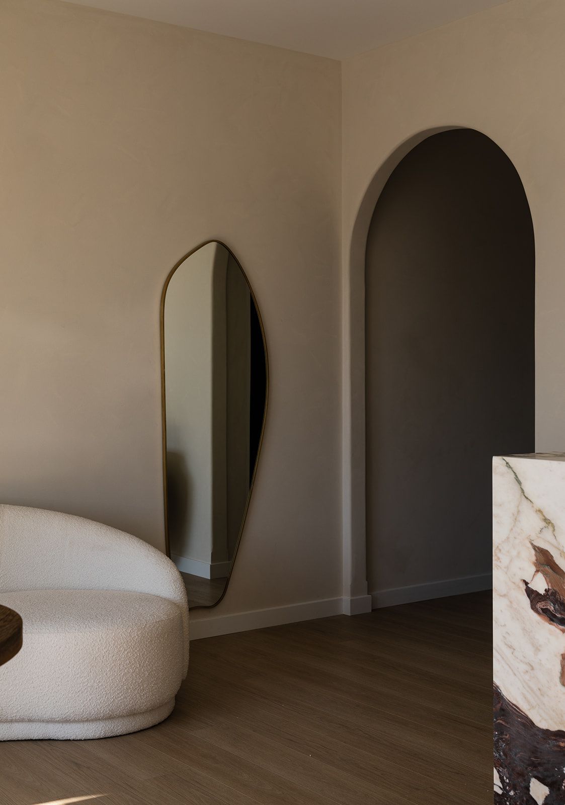 Modern minimalist interior with curved mirror, white curved sofa, and arched doorway against beige walls.