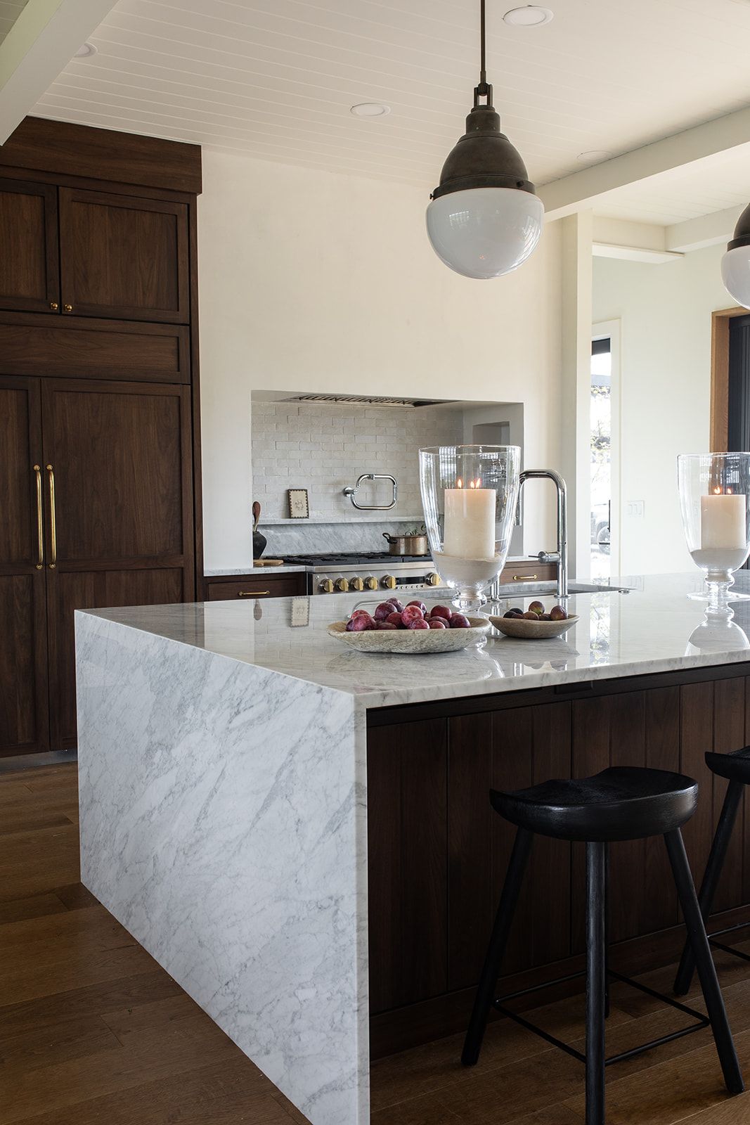 Luxury kitchen featuring dark wood cabinetry, thick marble waterfall island, black stools, candles, and industrial pendant lighting.