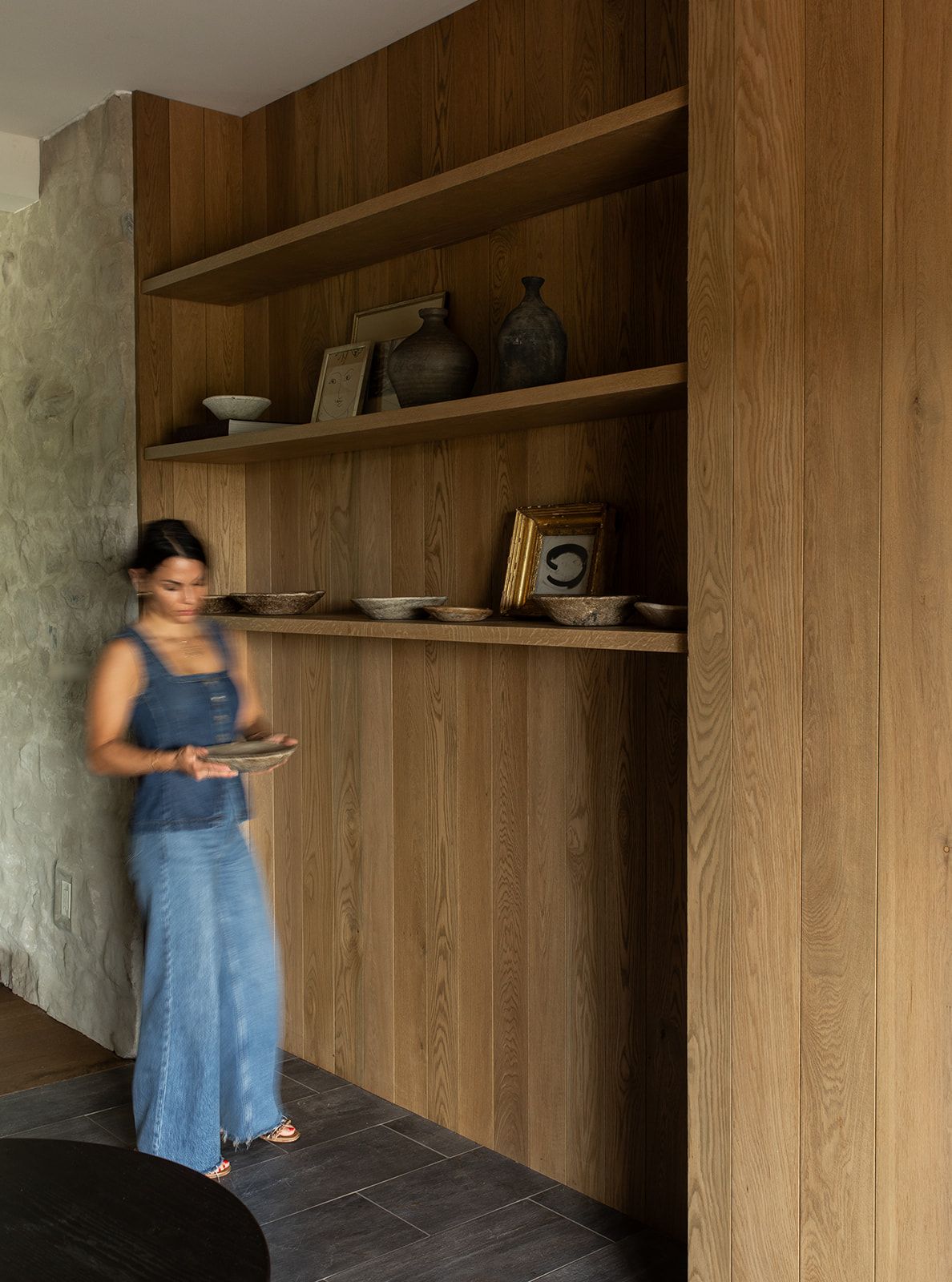 Woman browsing oak built-in shelves displaying ceramic vases, bowls, and decorative objects in a modern interior.