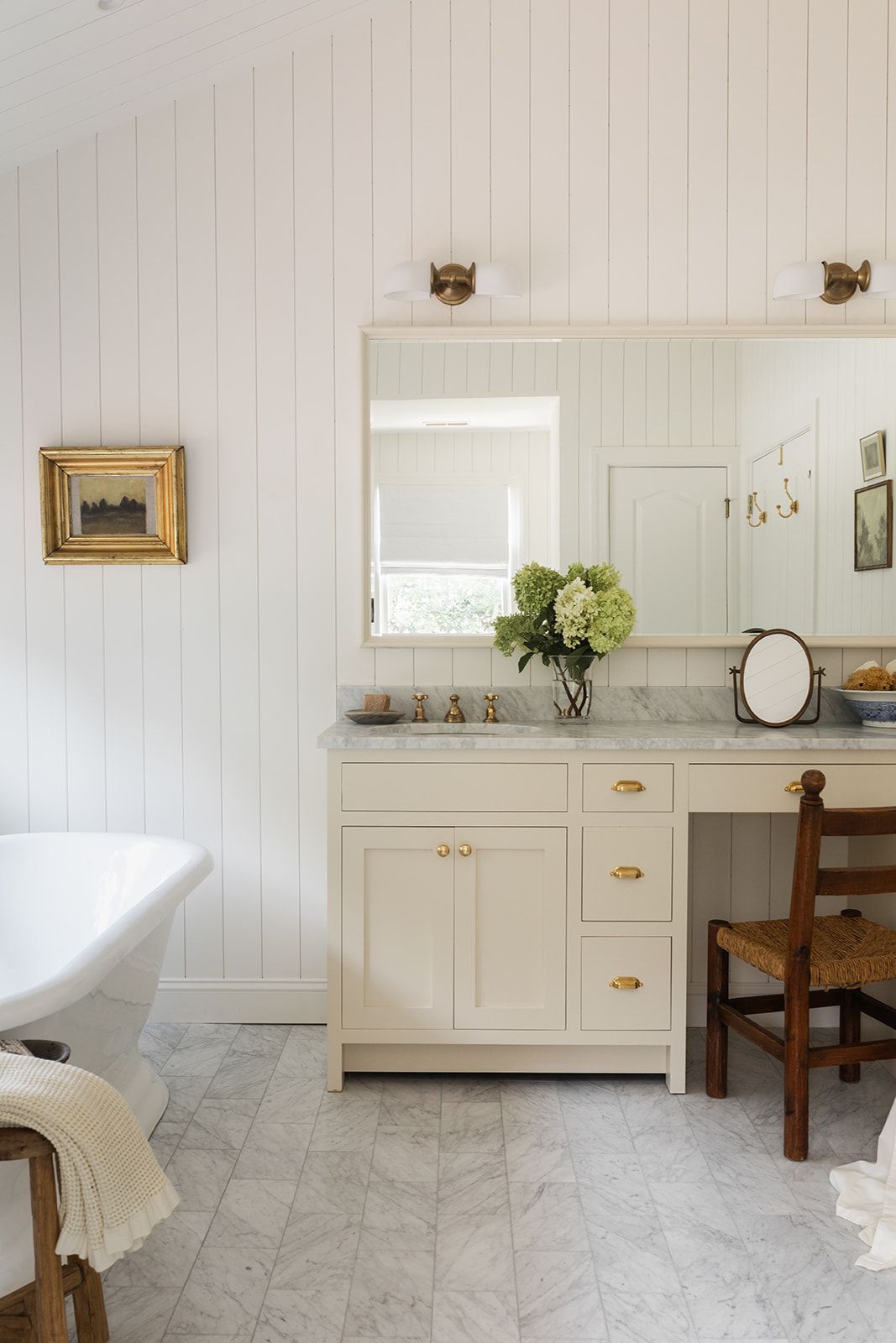 Elegant white bathroom with shiplap walls, marble floors, cream vanity with brass hardware, clawfoot tub, and fresh hydrangeas.
