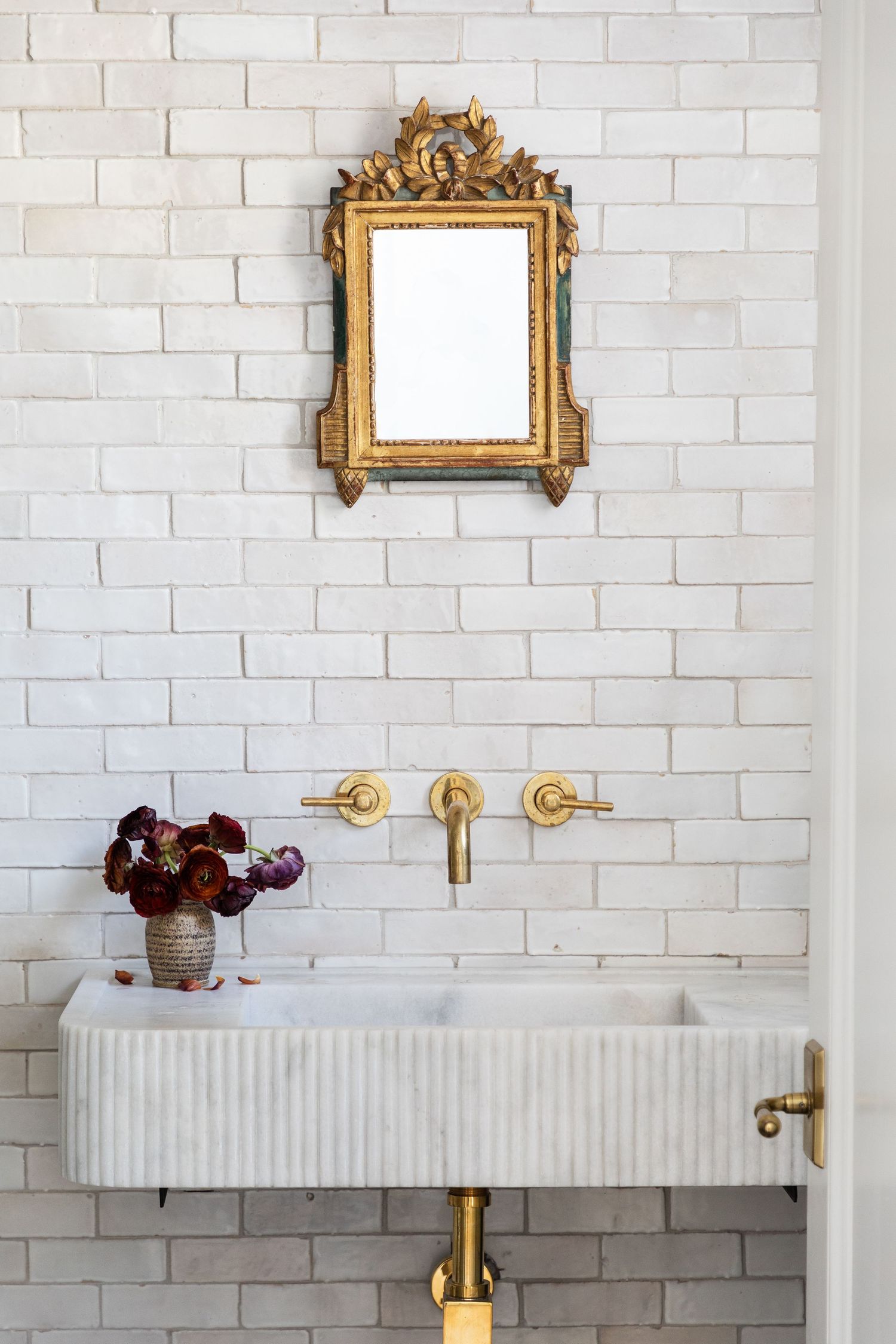 Elegant gold ornate mirror above white sink with brass wall-mounted faucet and purple flowers on white brick wall.