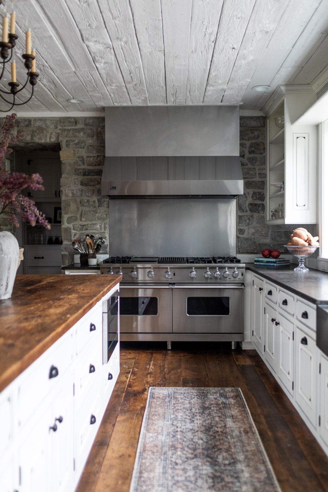 Modern rustic kitchen with white cabinets, stone walls, stainless steel appliances, butcher block counters and a vintage rug.