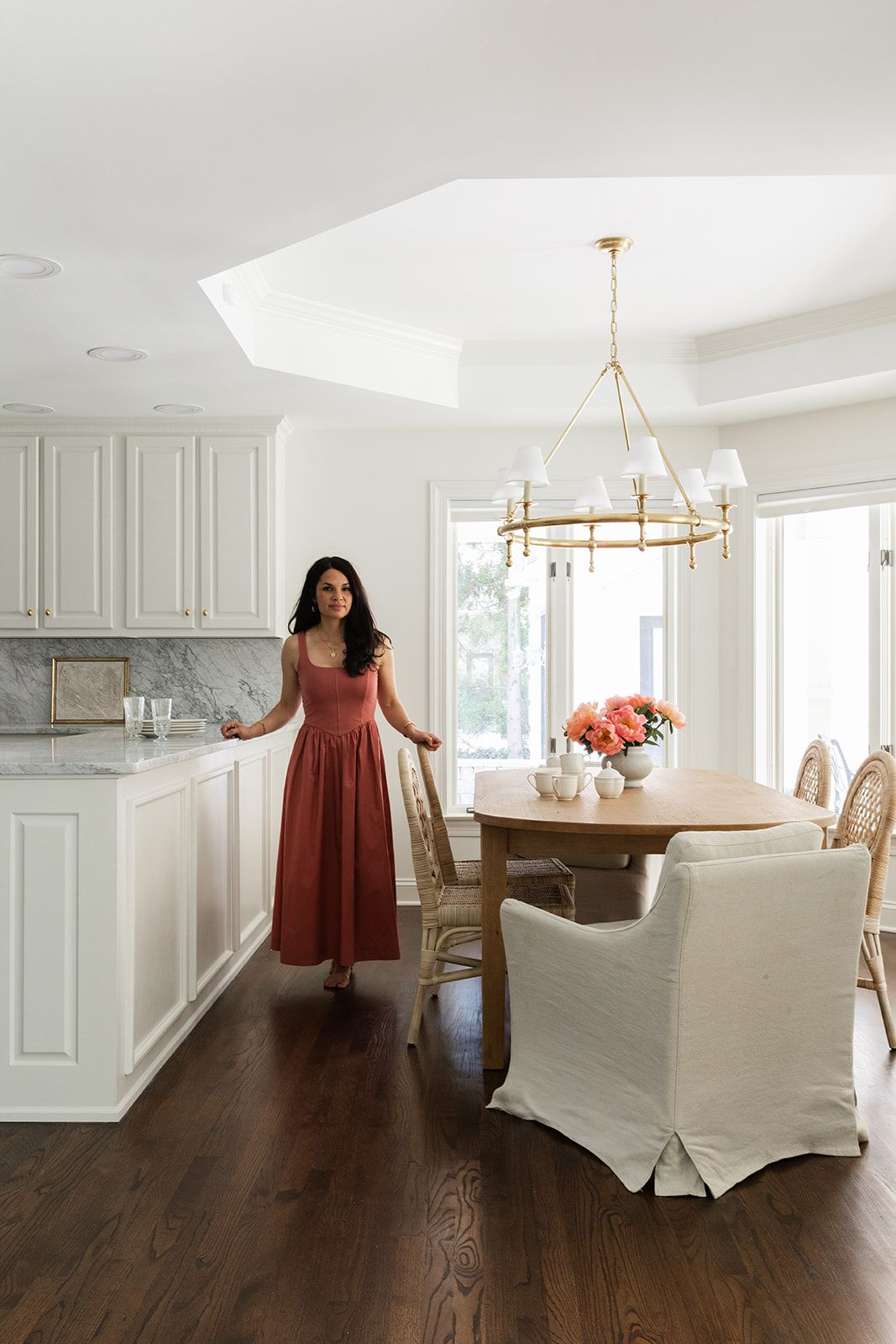 Woman in red dress standing in bright white kitchen with marble countertops and elegant dining area with gold chandelier.