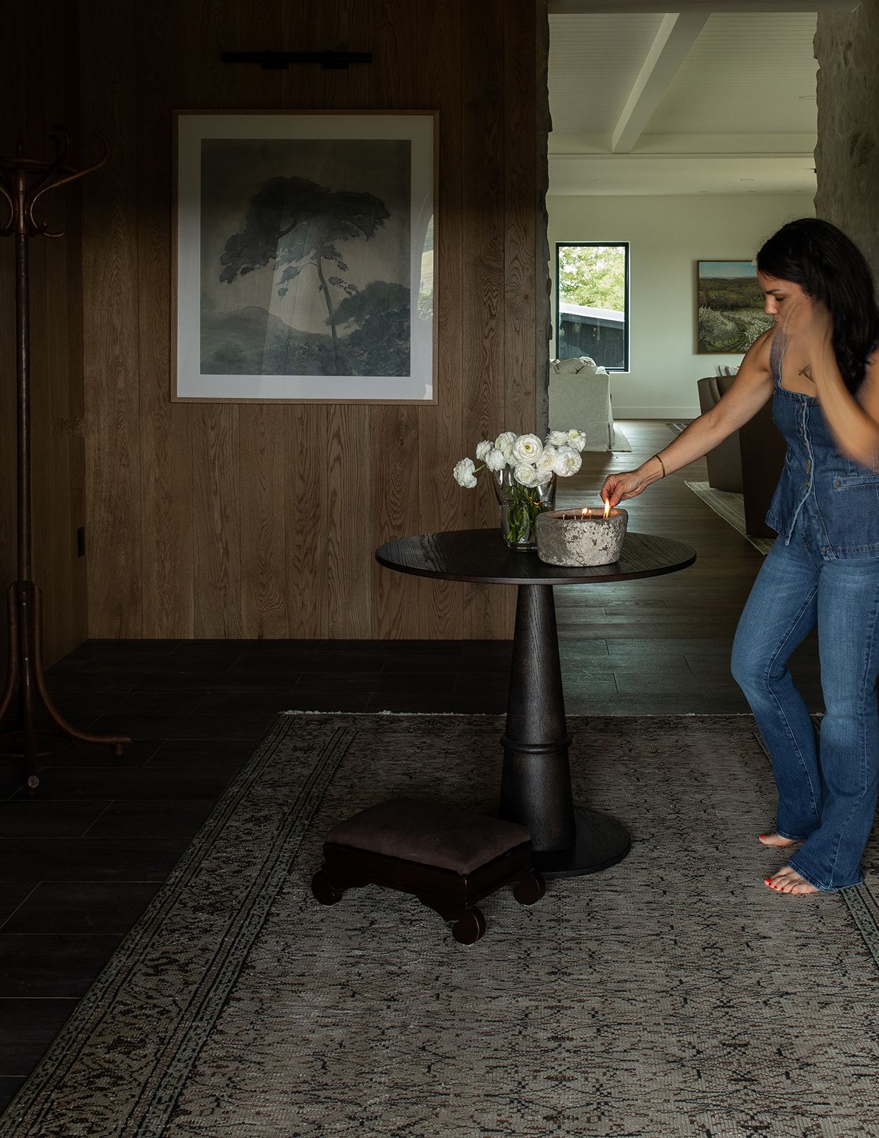 Woman lighting a candle on a dark pedestal table with white flowers, vintage rug, and framed art in a modern home.