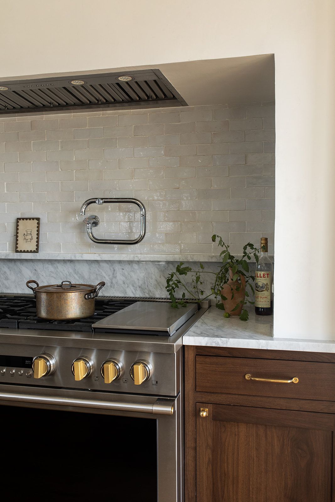 Close-up of professional stainless range with brass knobs, gray zellige tile backsplash, and dark walnut cabinetry.