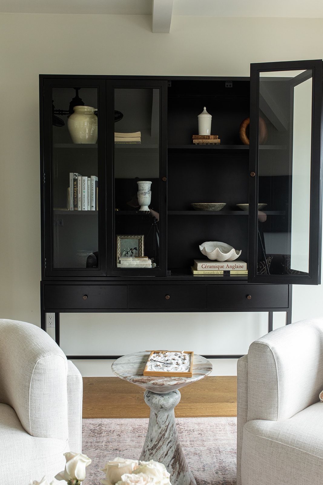 Black glass-front display cabinet with ceramic decor, flanked by two white upholstered armchairs and marble side table.