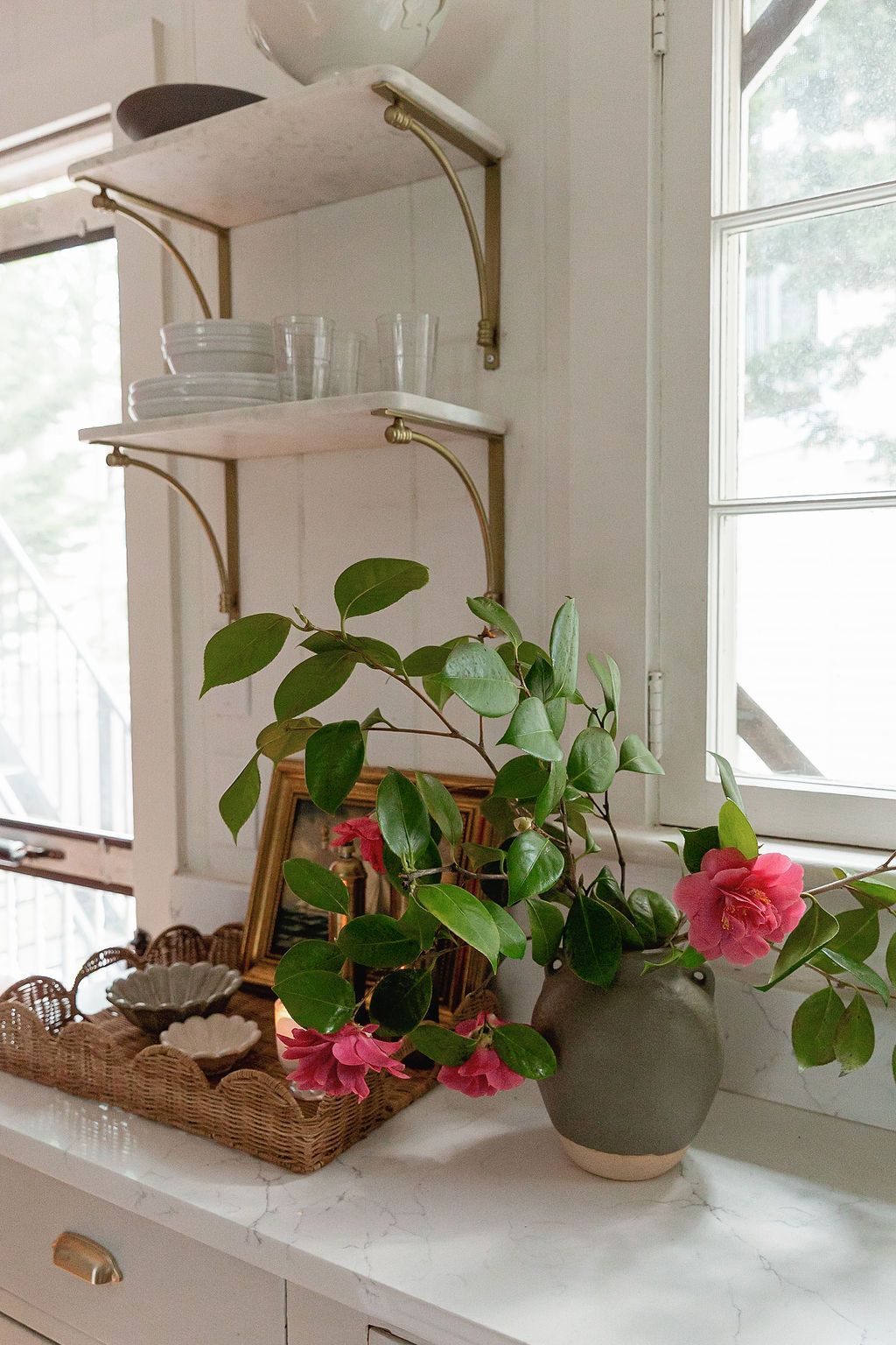 Vintage-style shelving displays glass cups above a windowsill with pink flowering plants in a ceramic pot.