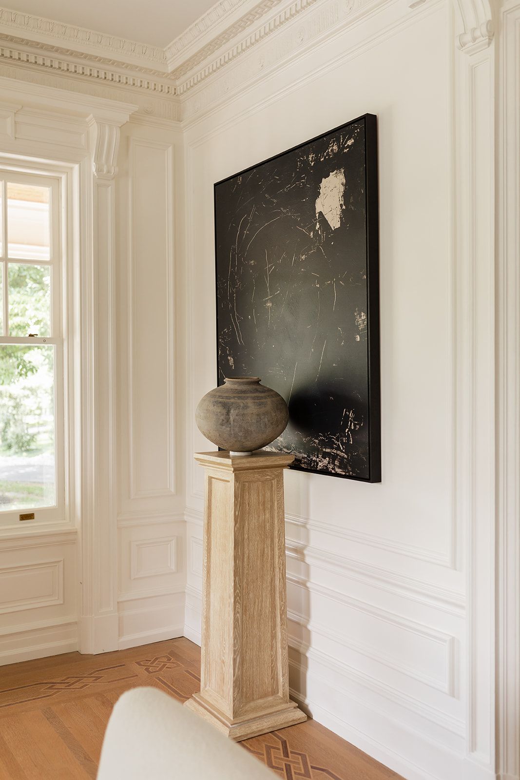 White paneled room with crown molding features dark artwork and a wooden pedestal against pristine walls.