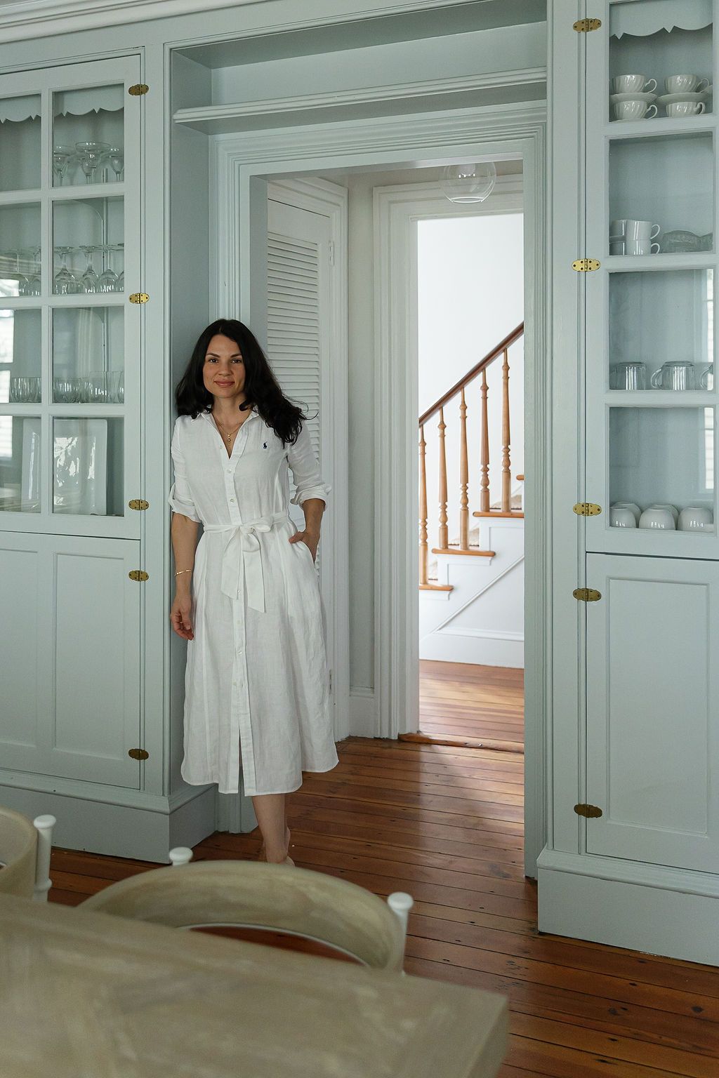 Figure in white dress stands beside mint green built-in cabinets in elegant living space with hardwood floors.
