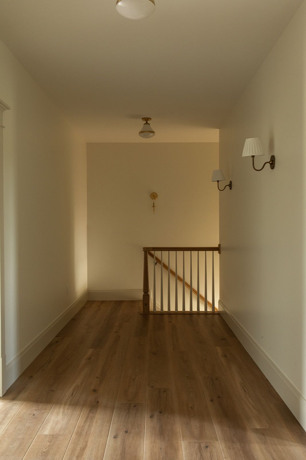 Bright hallway with wooden floors, white walls, wall sconce, and wooden staircase railing.