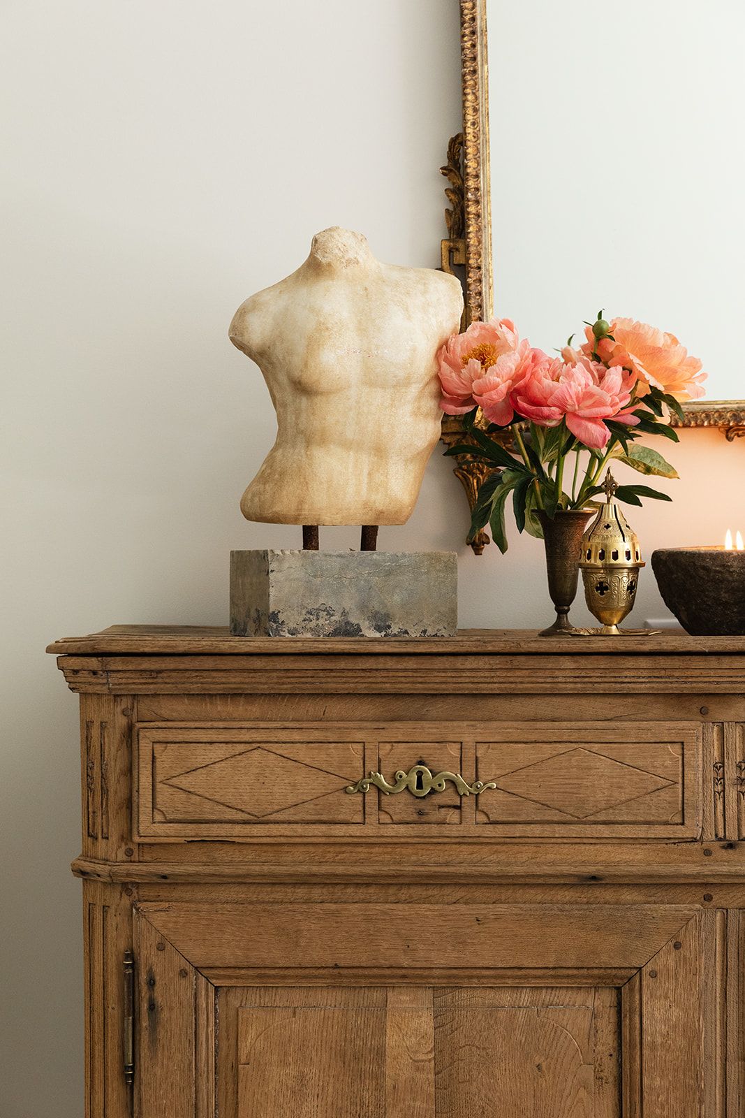 Antique wooden dresser with marble torso sculpture, pink peonies in gold vase, and ornate mirror on neutral wall.