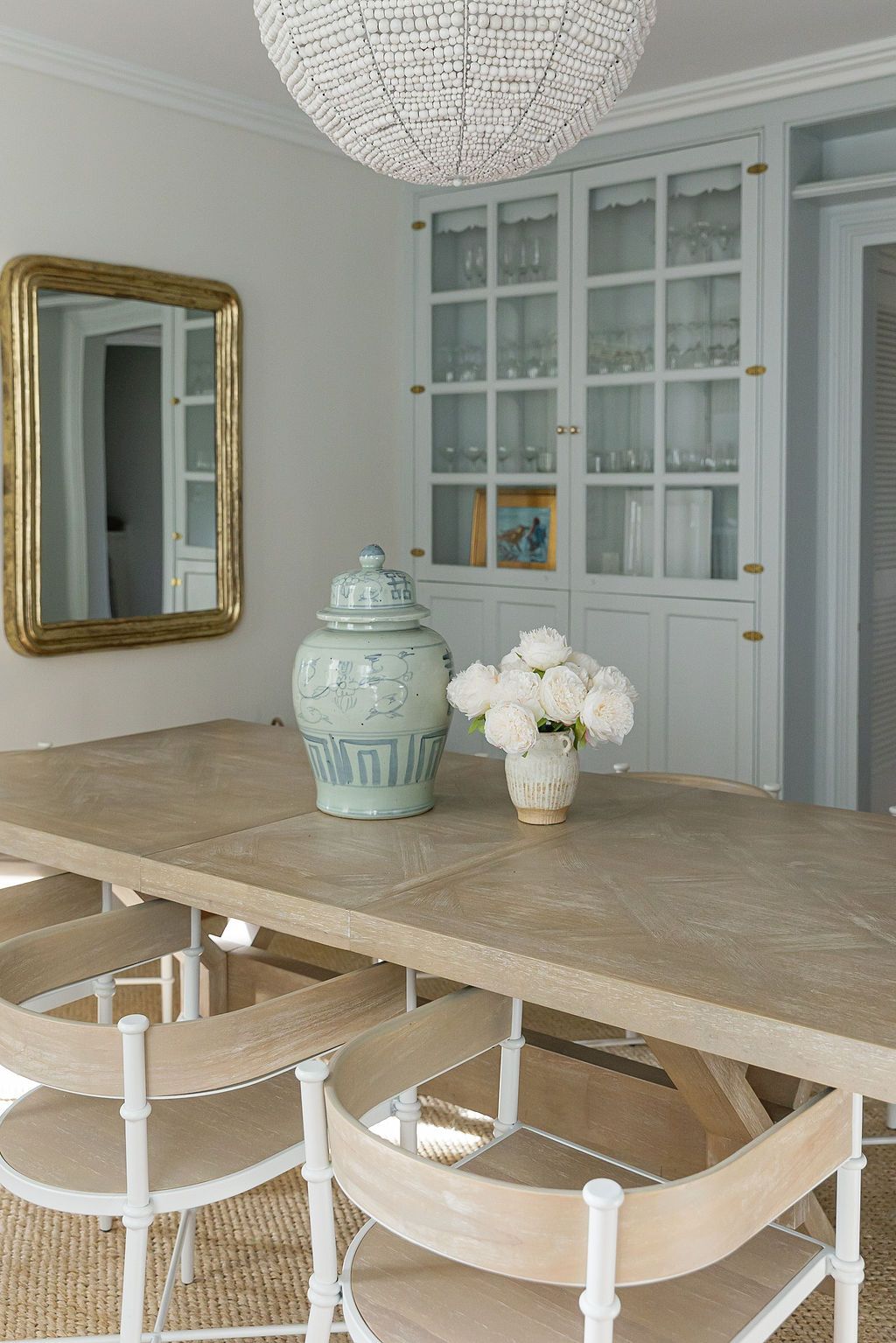 Elegant dining room with blue-gray built-ins, crystal chandelier, gold mirror, and light wood dining set on sisal rug.