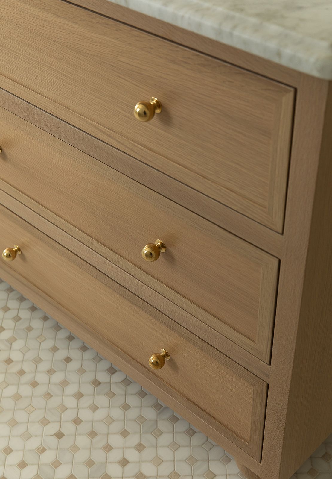 Close-up of a light oak wood dresser with three drawers and round brass gold knobs on a mosaic tile floor.
