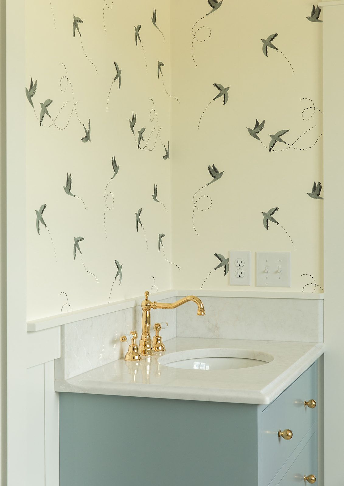 Close-up of sage green bathroom vanity with brass faucet, marble countertop, and whimsical bird-patterned wallpaper.