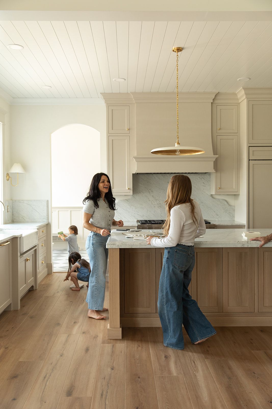Two women chat in a bright modern kitchen with shiplap ceiling, marble backsplash, and children playing nearby.
