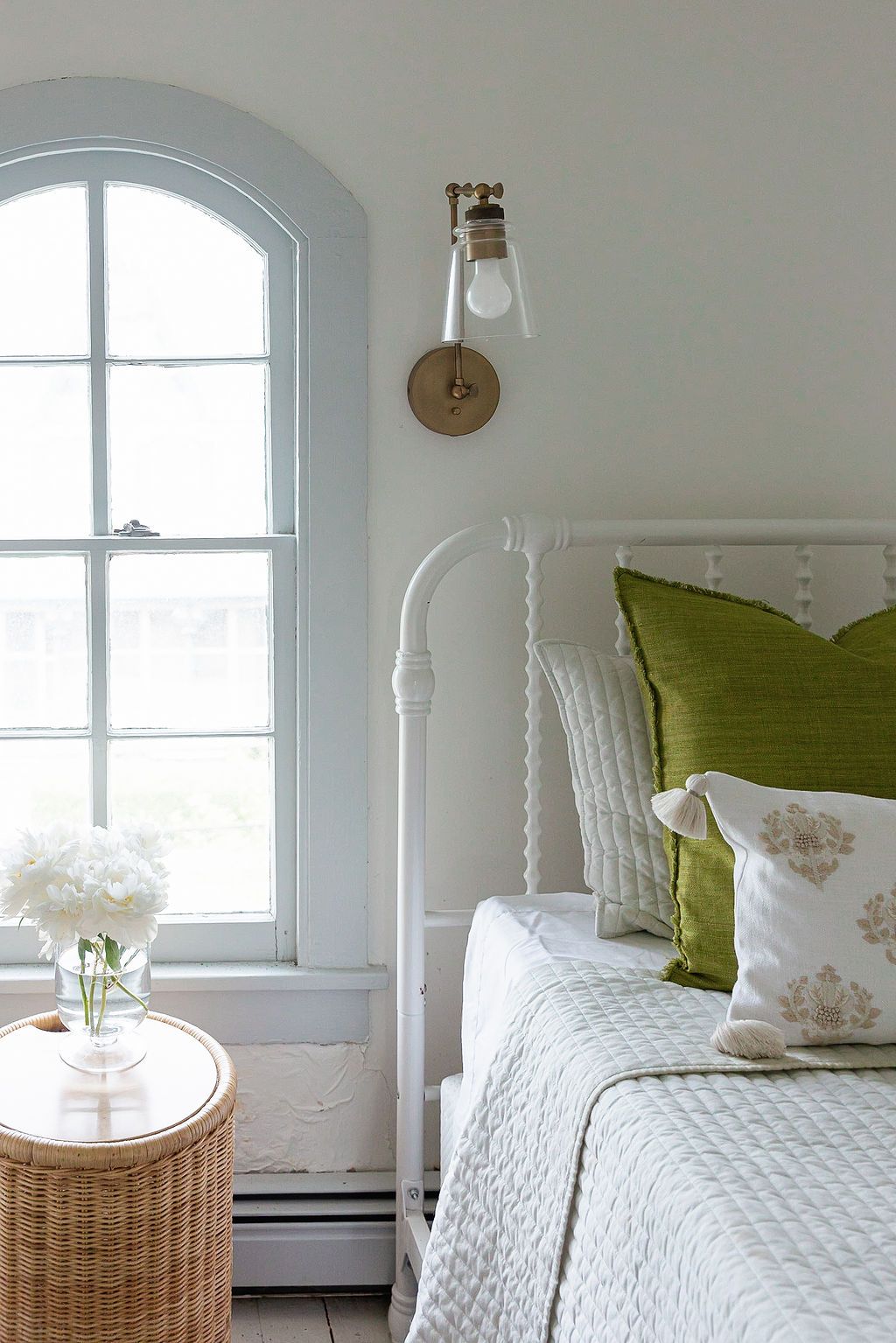Serene white bedroom with green accent pillows features a quilted bedspread, arched window, and woven rattan side table.