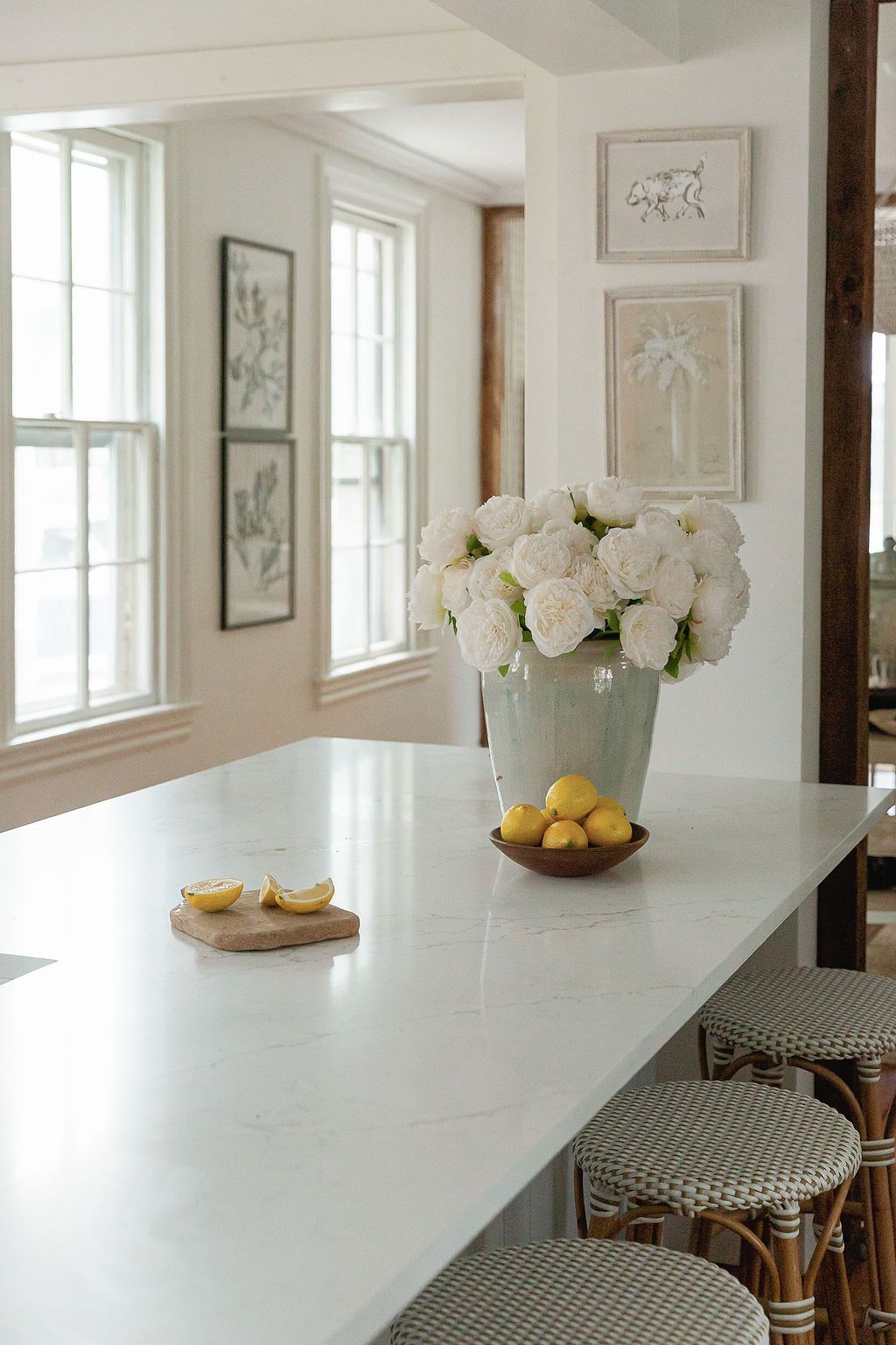 Bright kitchen counter with white roses in a metal vase, lemons on a wooden board, and bistro chairs with checkered seats.