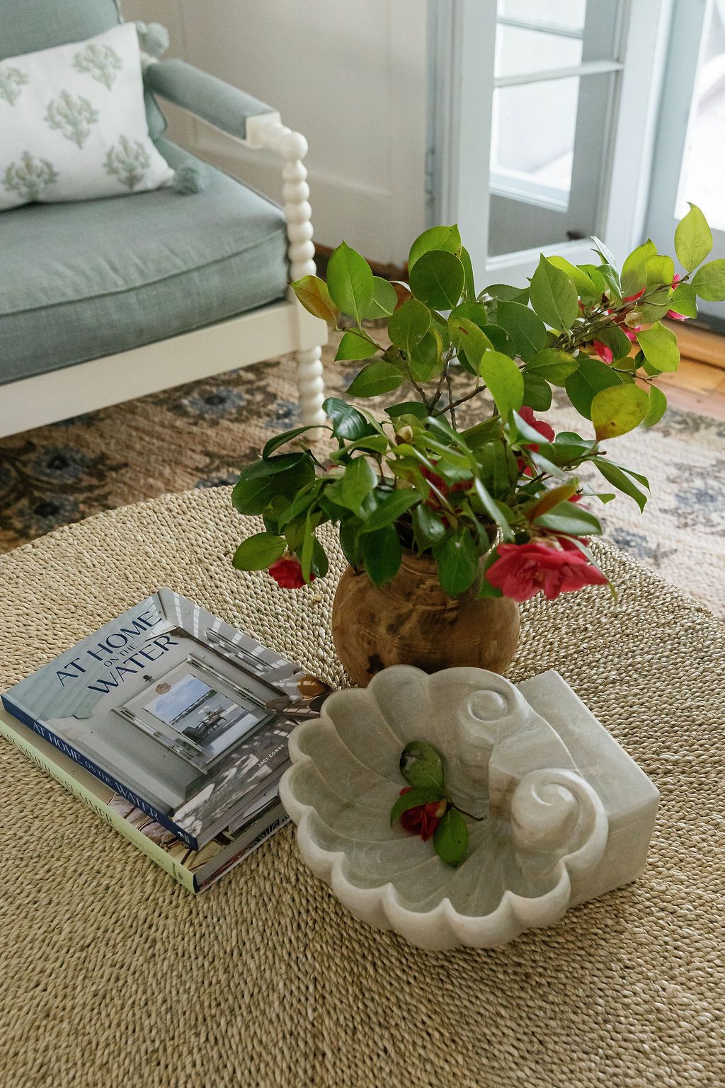 A cozy living room corner with a sage green sofa, potted red flowers, decorative bowl, and magazines on a jute rug.