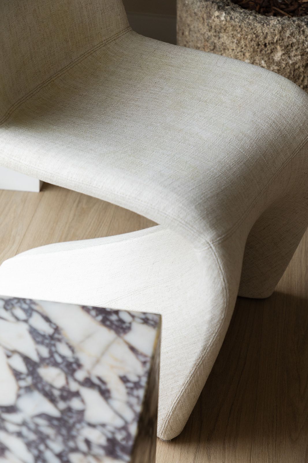 Close-up of a curved modern upholstered chair in off-white linen fabric with marble side table on wooden floor.