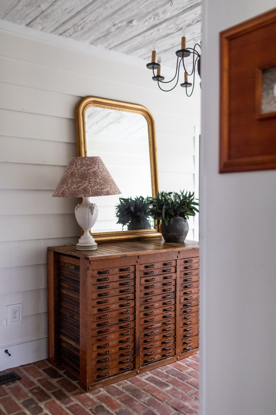Vintage wooden cabinet with multiple drawers topped with a gold-framed mirror, table lamp and plants on brick flooring.