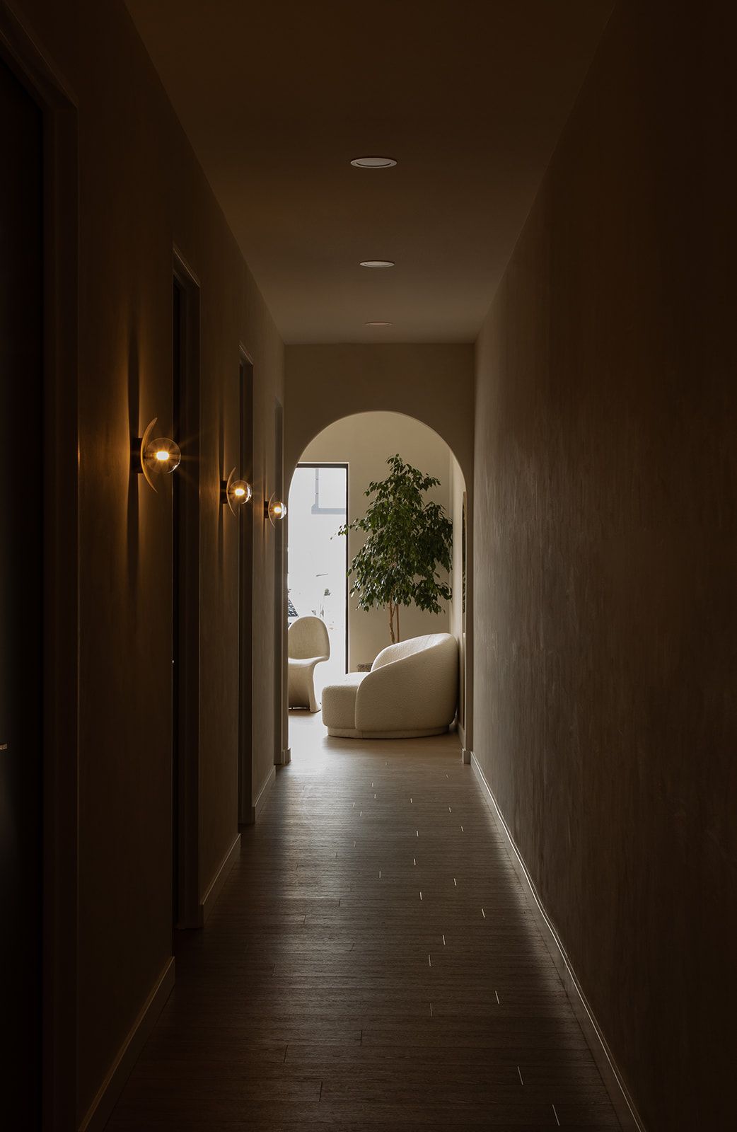Dark hallway with wall sconces leads to a bright room with an arched doorway, white furniture and a potted plant.