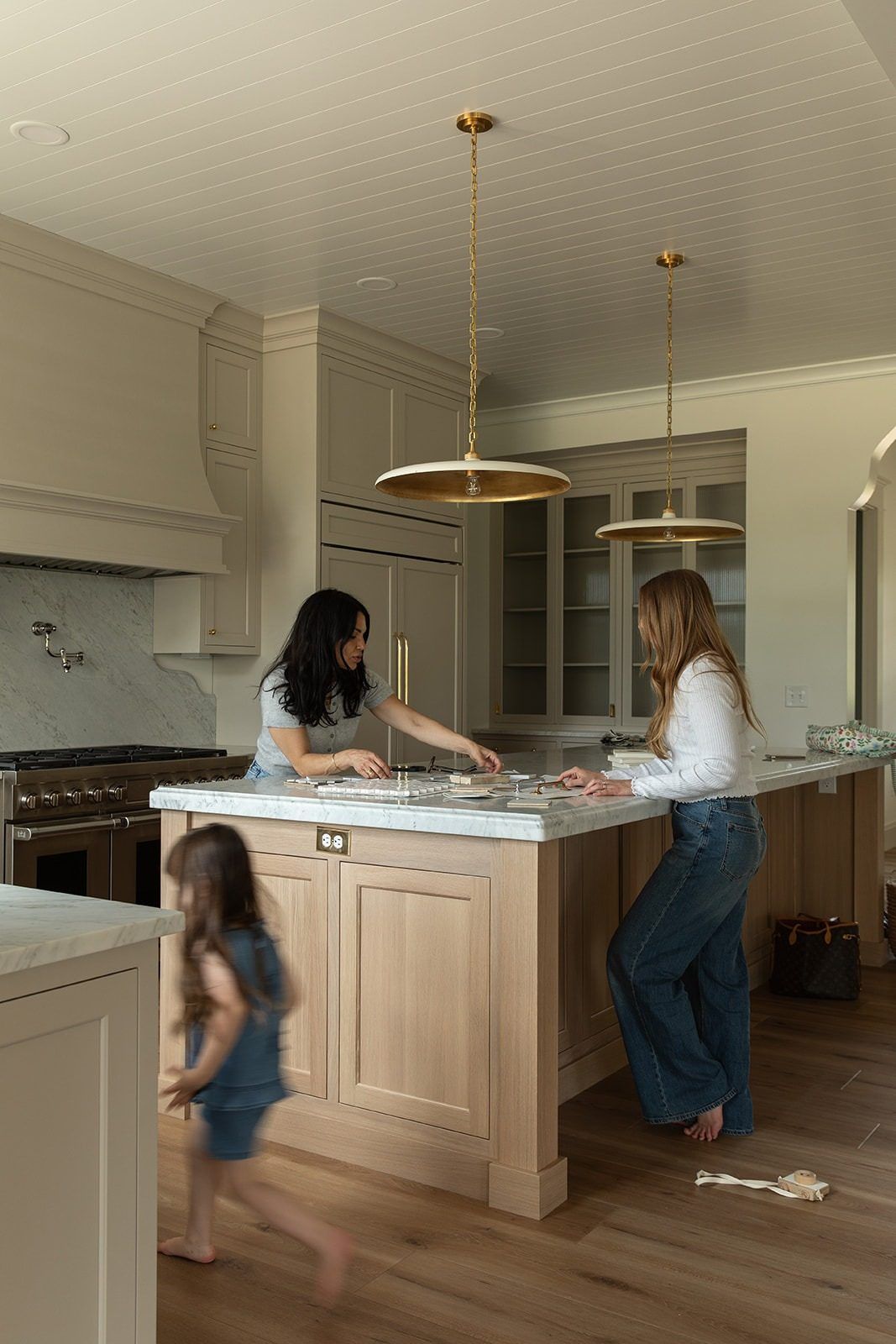 Two women work at a marble kitchen island while a child runs past in a neutral-toned kitchen with brass pendants.