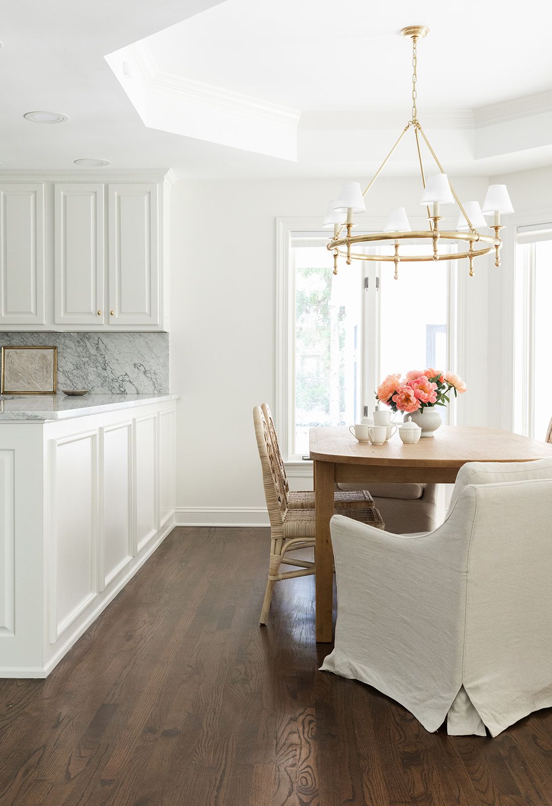 Elegant white dining room featuring gold ring chandelier, oval wood table, slipcovered chair, rattan seats, and marble kitchen counter.