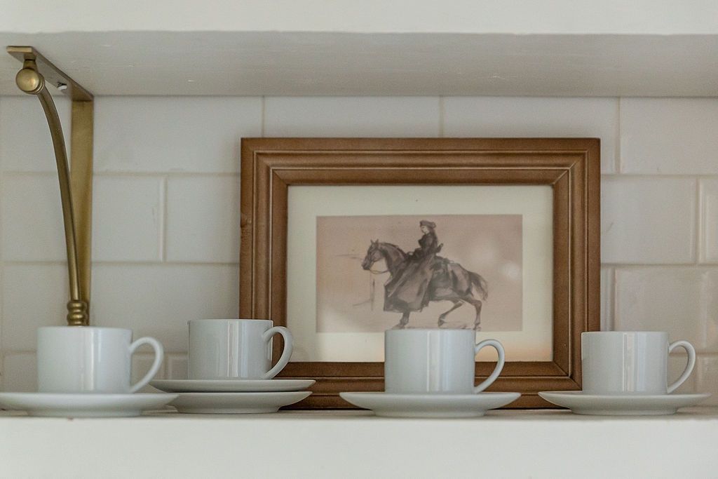 Four white coffee cups and saucers arranged on a shelf with vintage equestrian artwork framed in the background.