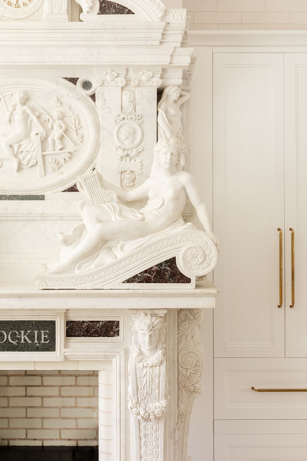 Ornate white marble fireplace mantel with detailed classical sculptural elements and brass cabinet hardware.
