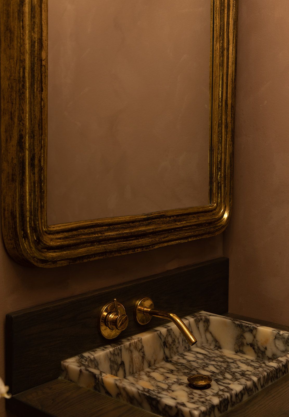 A vintage gold-framed mirror hangs above an elegant marble sink with brass wall-mounted faucet in a dimly lit bathroom.