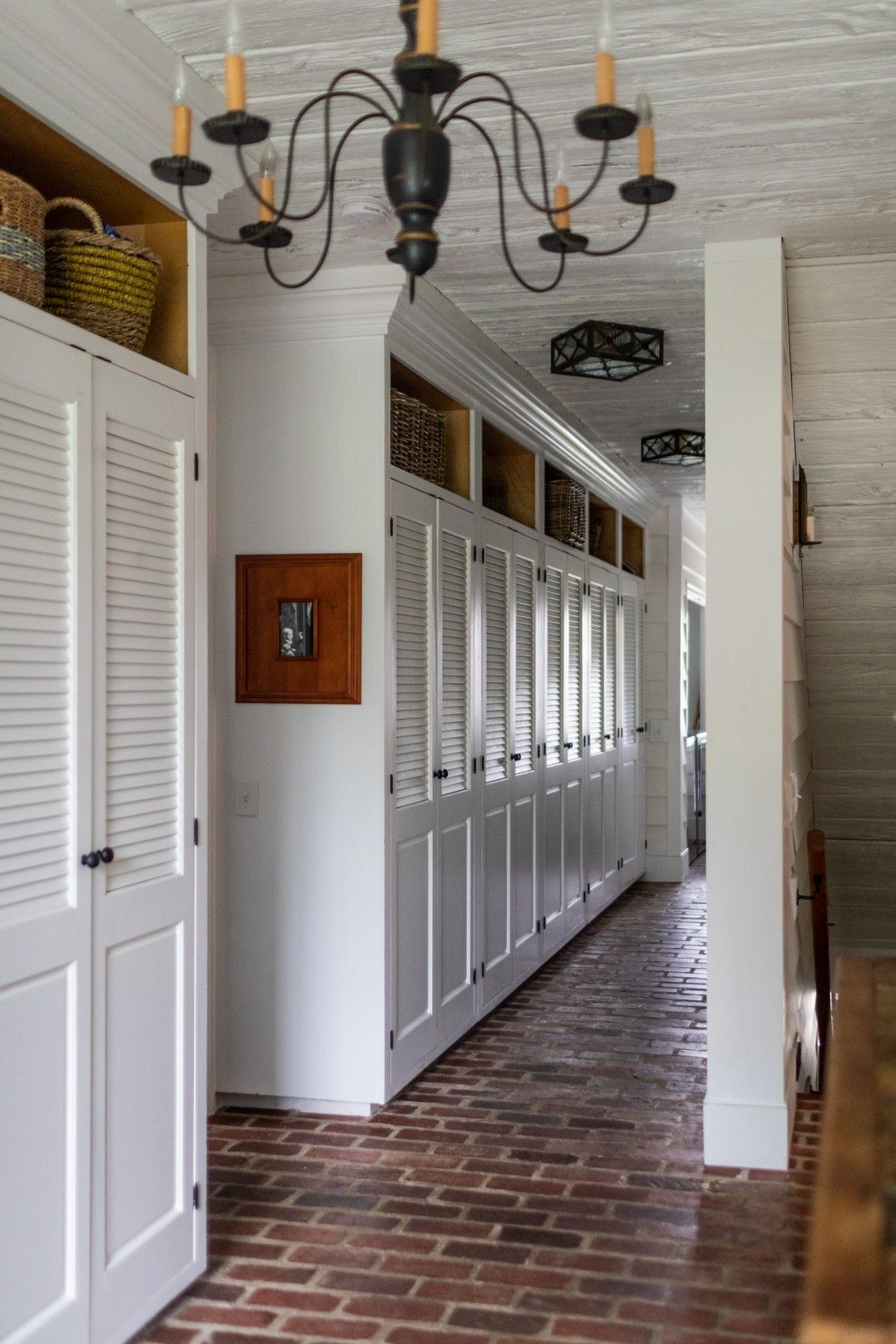 A long corridor with white louvered doors, brick flooring, and a black iron chandelier features built-in storage above.