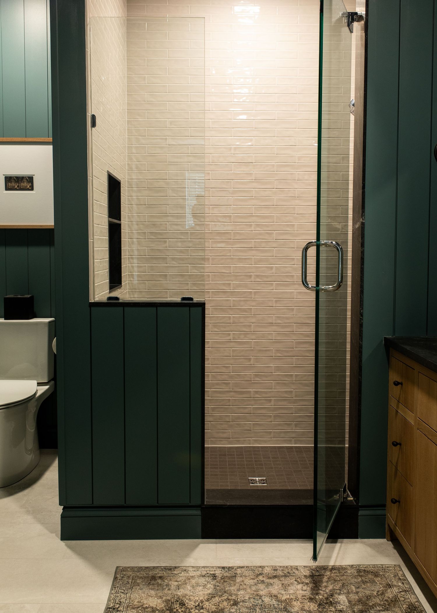 Modern bathroom interior with dark green wainscoting, glass shower door, and decorative area rug on white tile floor.
