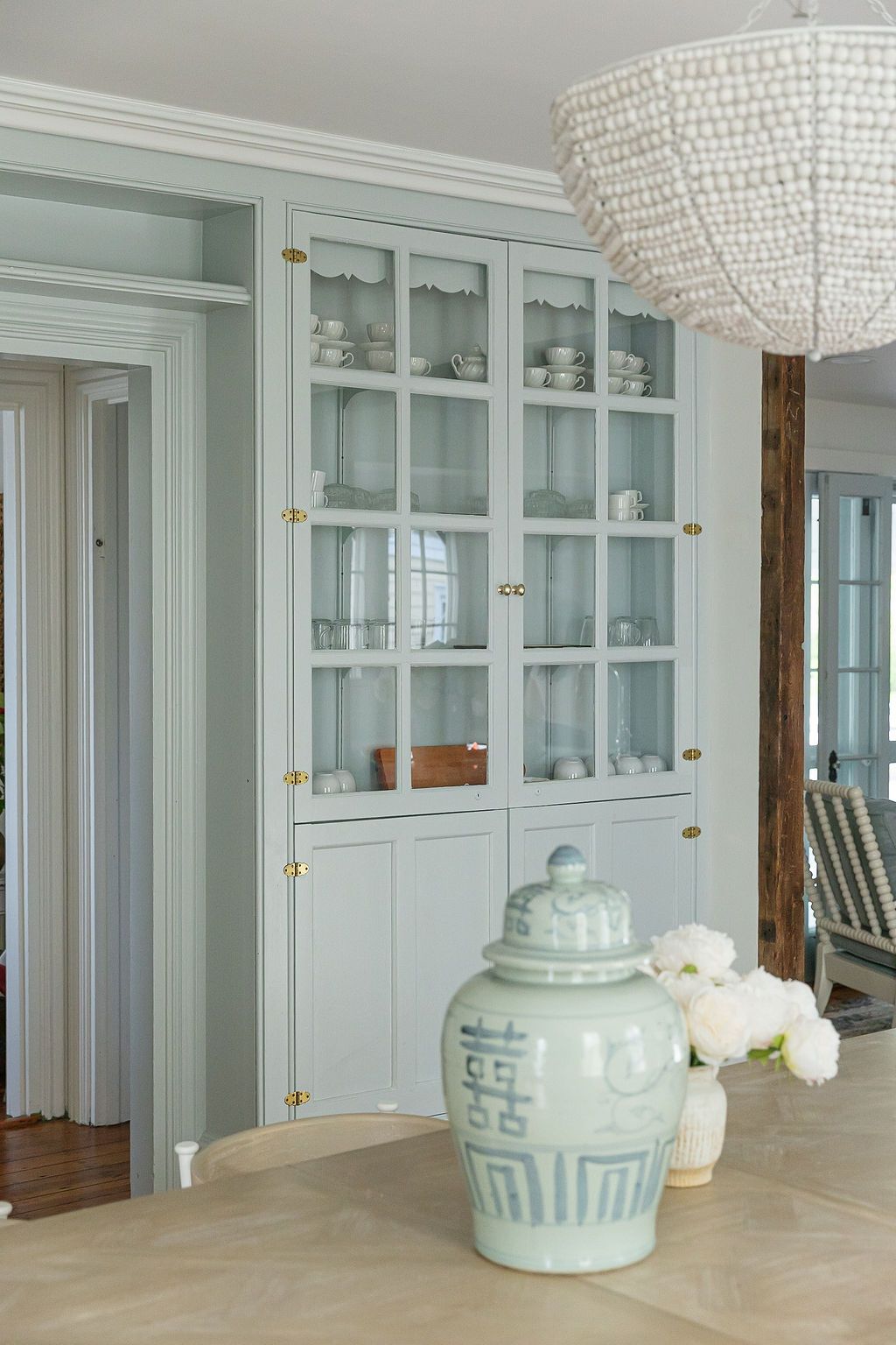 Elegant built-in glass cabinet painted in soft mint green showcases white dishes below a beaded chandelier.
