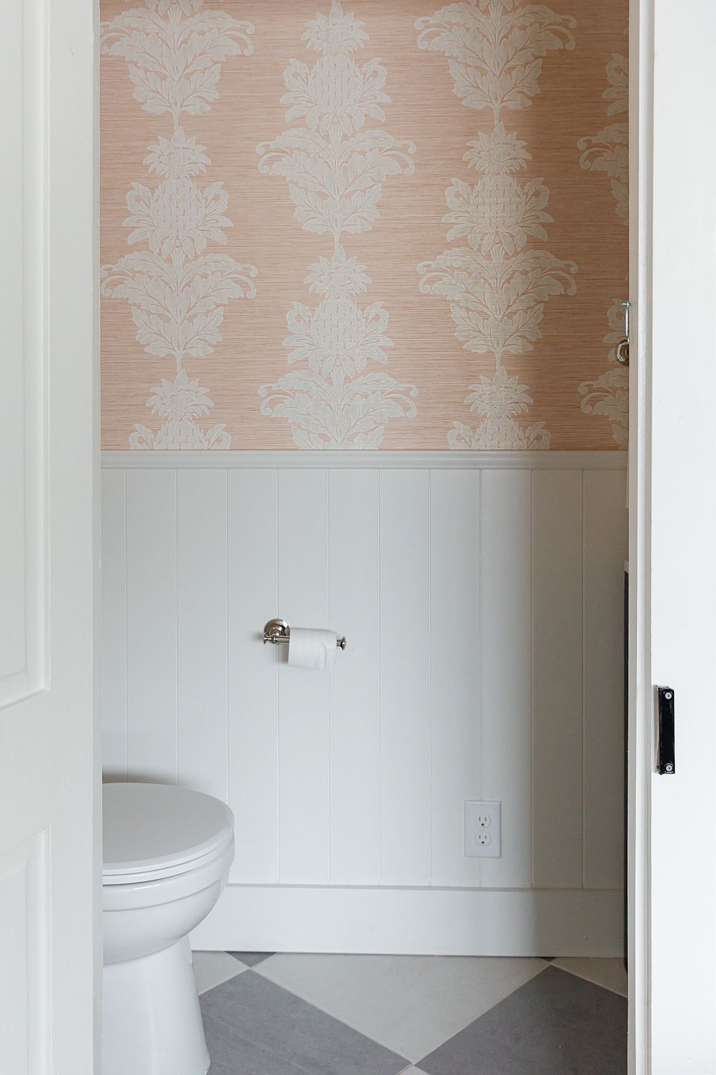 A powder room featuring blush pink damask wallpaper, white wainscoting and gray tile flooring.