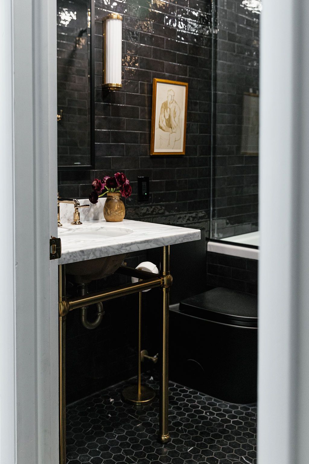 Modern bathroom with black tile walls, marble sink vanity, brass fixtures, hexagon floor tiles, and framed artwork.