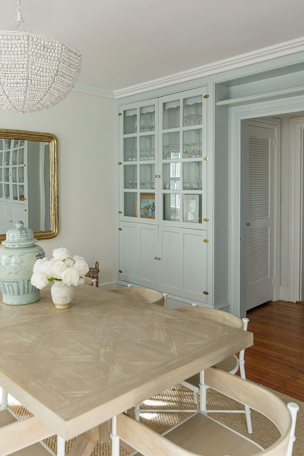 A coastal-style dining room features a crystal chandelier, built-in glass cabinets, and a light-colored herringbone table.