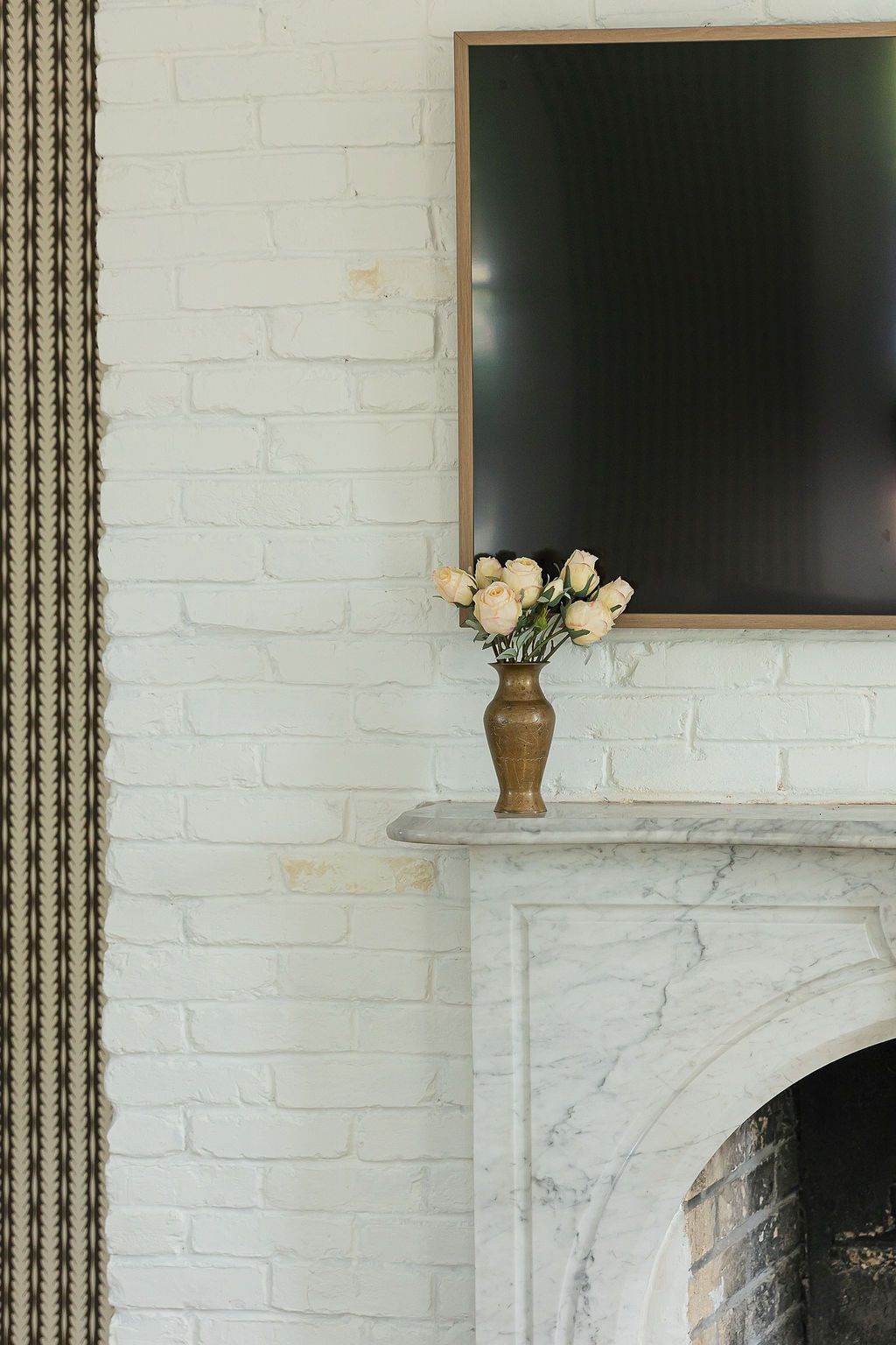 White vase with cream roses sits atop a marble fireplace mantel against white brick wall with dark frame above.