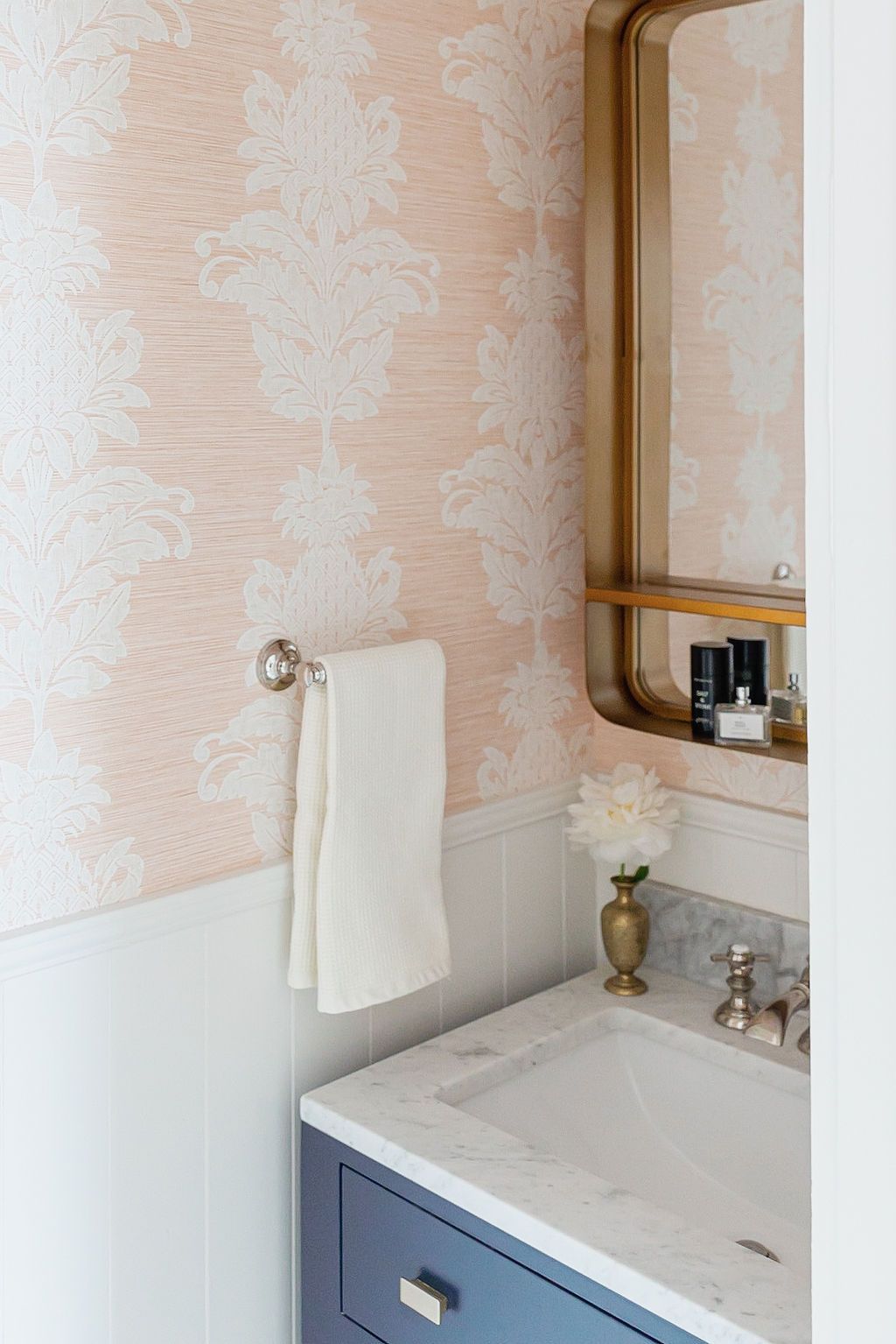 A bathroom vanity with blue cabinets and marble top against blush damask wallpaper features a brass mirror and white towel.