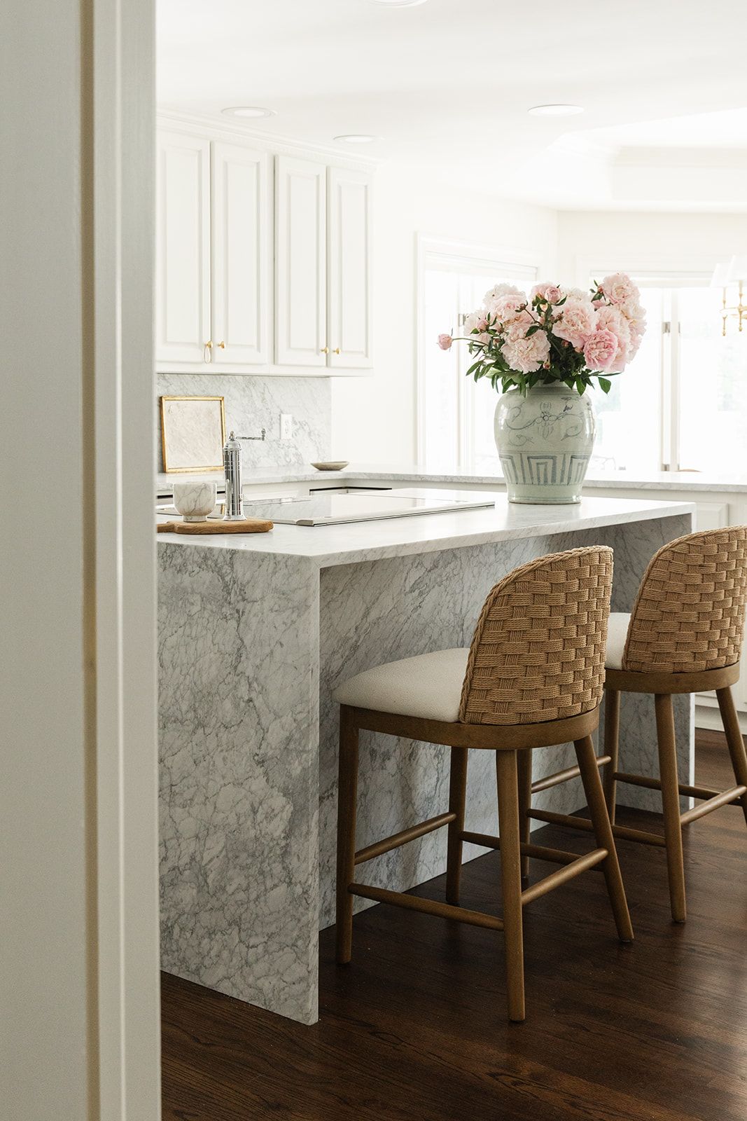 White kitchen with marble island, woven counter stools, and pink peonies in a vase on dark hardwood floors.