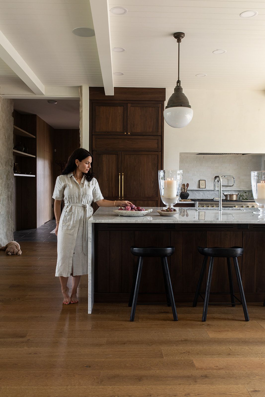 Woman in white robe standing at dark wood kitchen island with marble countertop, black stools, and pendant light.