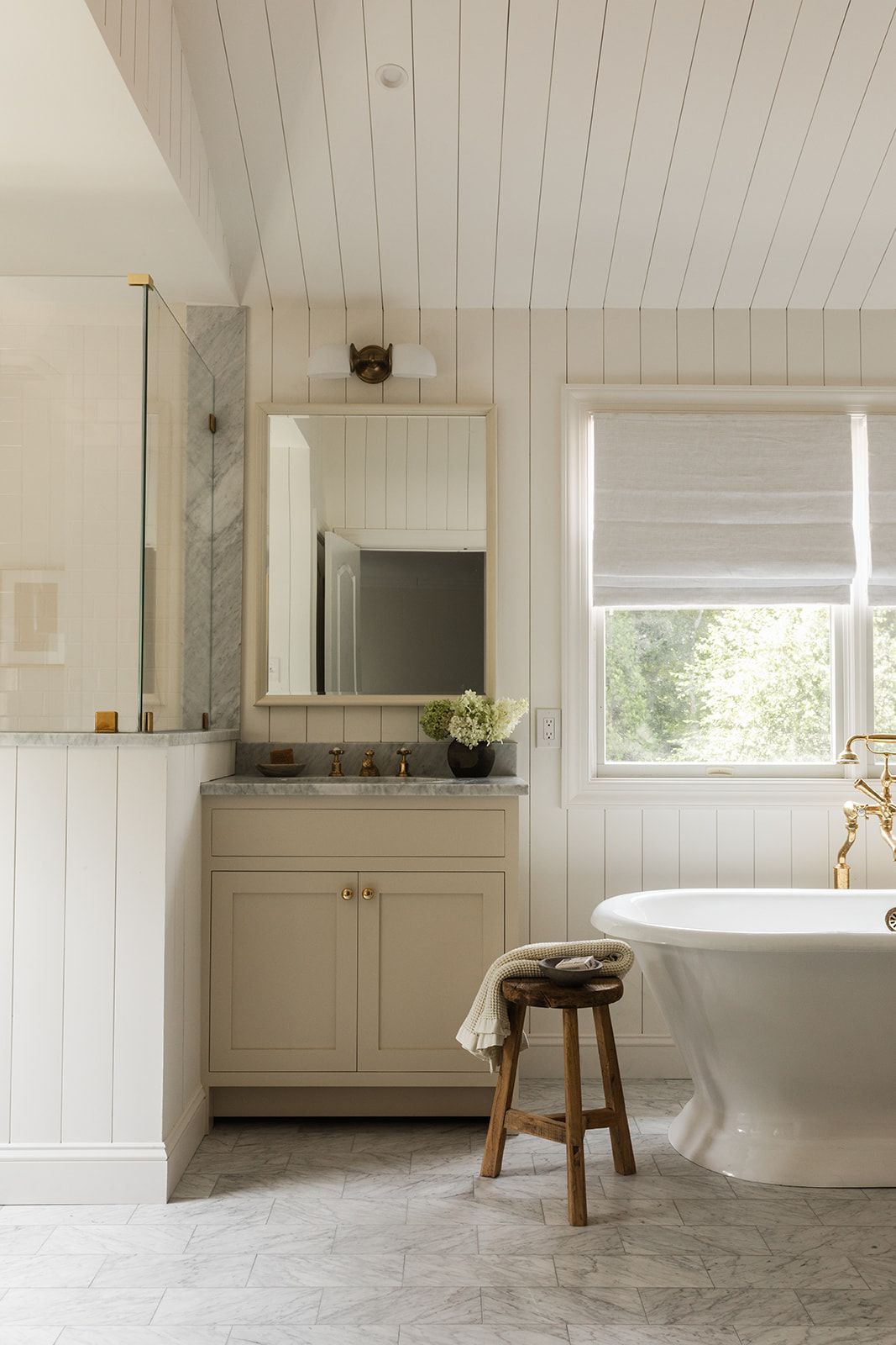 Elegant white shiplap bathroom featuring cream cabinets, marble accents, freestanding tub, brass faucet, and wooden stool.